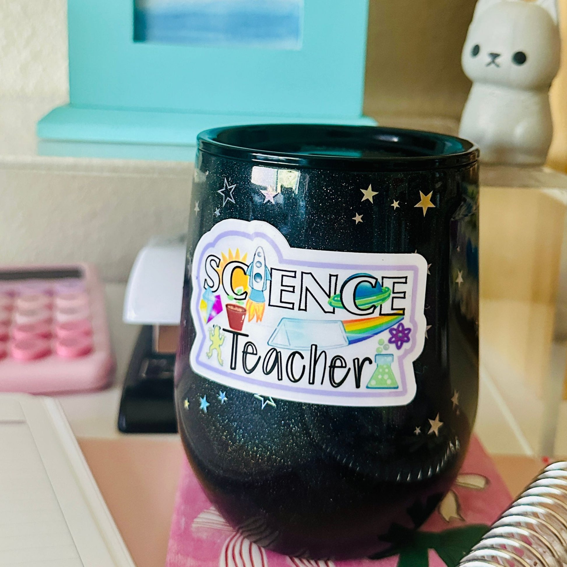 A black glitter stemless wine tumbler with a colorful 'Science Teacher' sticker on the front, set on a desk with a pink calculator and a white cat figurine in the background.