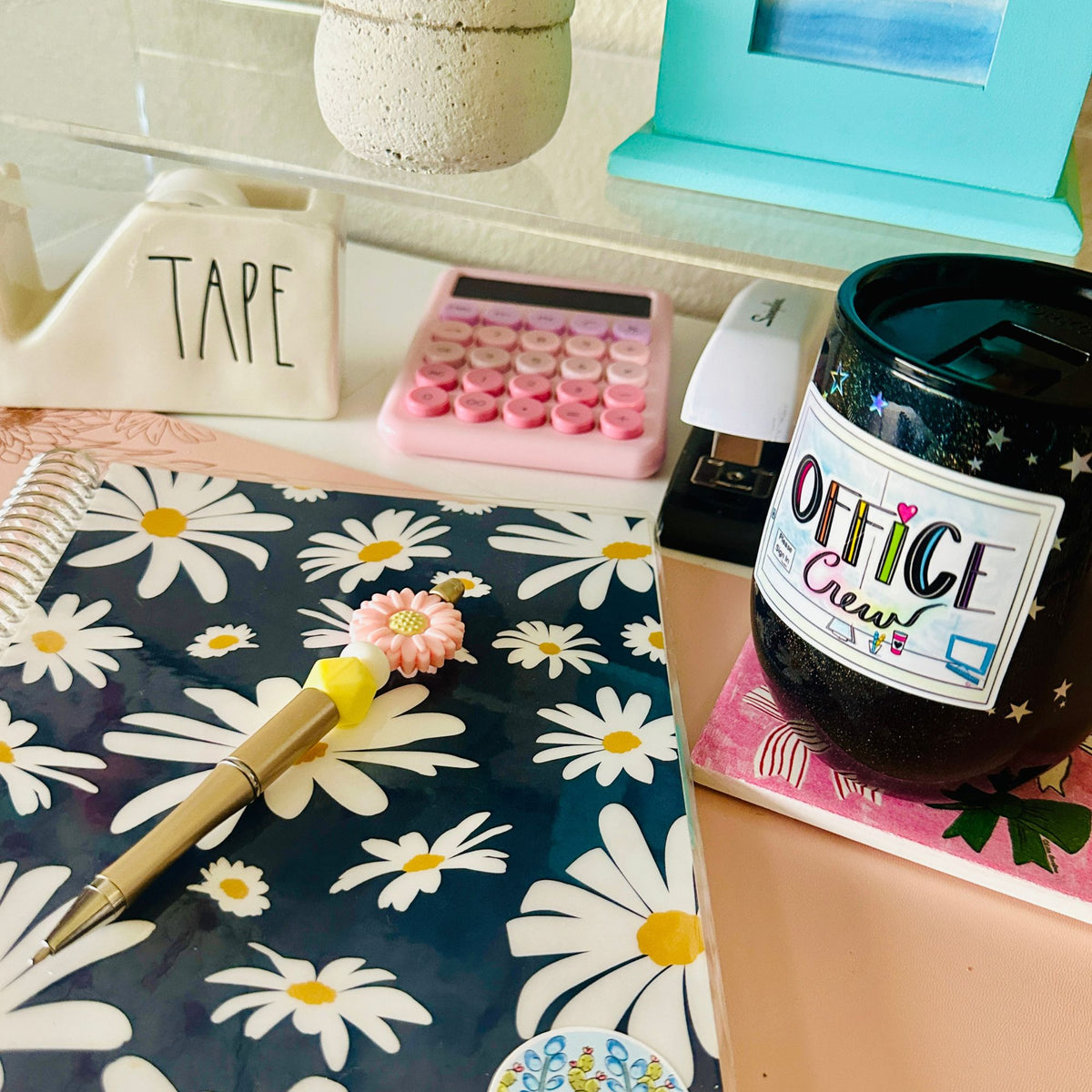 A styled desk scene featuring a navy blue notebook with a white daisy pattern, a gold pen, a pink flower-shaped clip, a pink calculator, and a black ceramic mug with 'OFFICE Crew' printed on it.