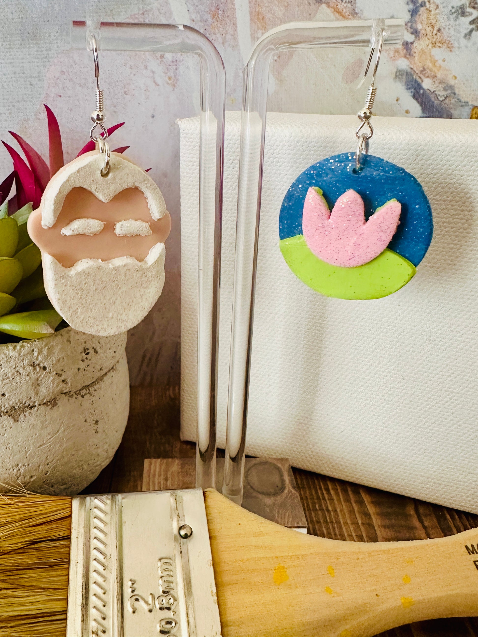Close up of handmade Claude Monet portrait and water lily earrings displayed with art supplies and colorful backdrop.