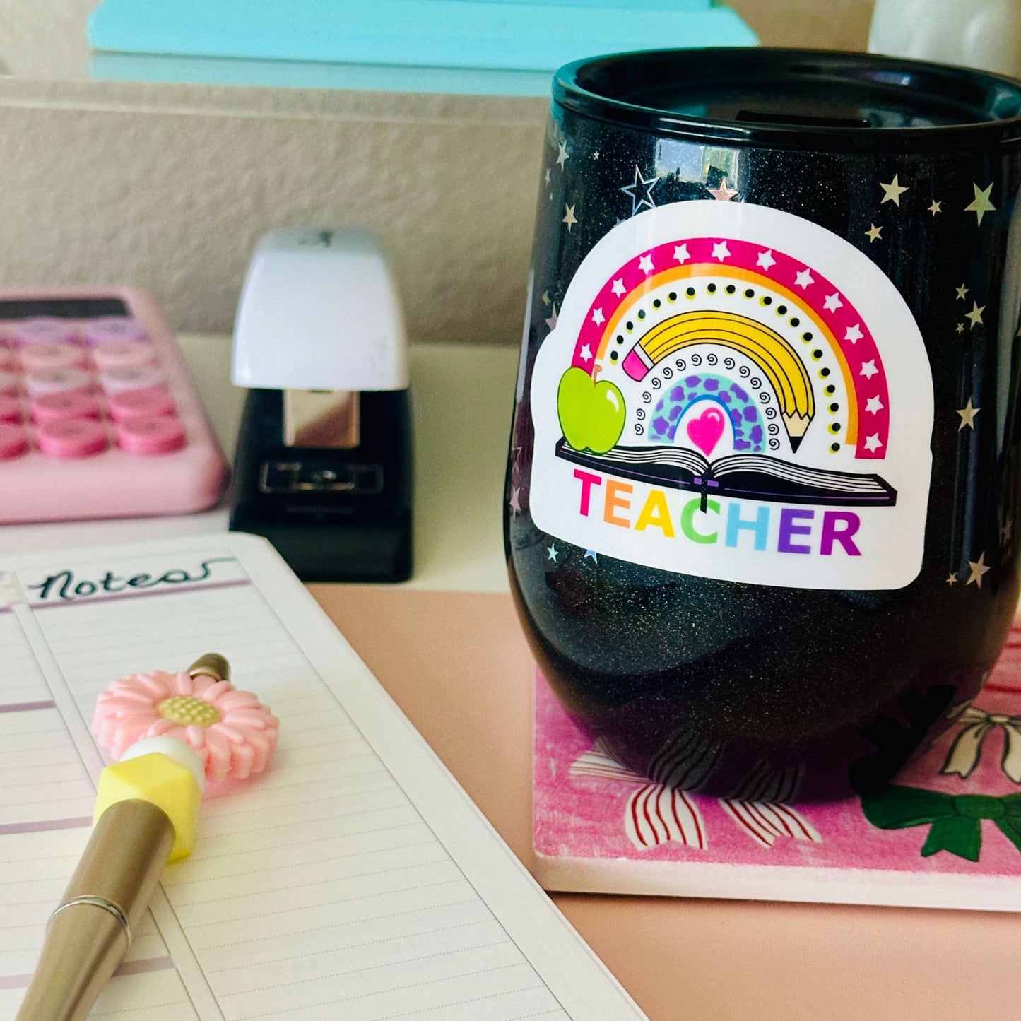 A black glitter tumbler cup with a colorful rainbow and apple 'TEACHER' sticker, styled on a desk next to a notebook, pen, calculator, and stapler.