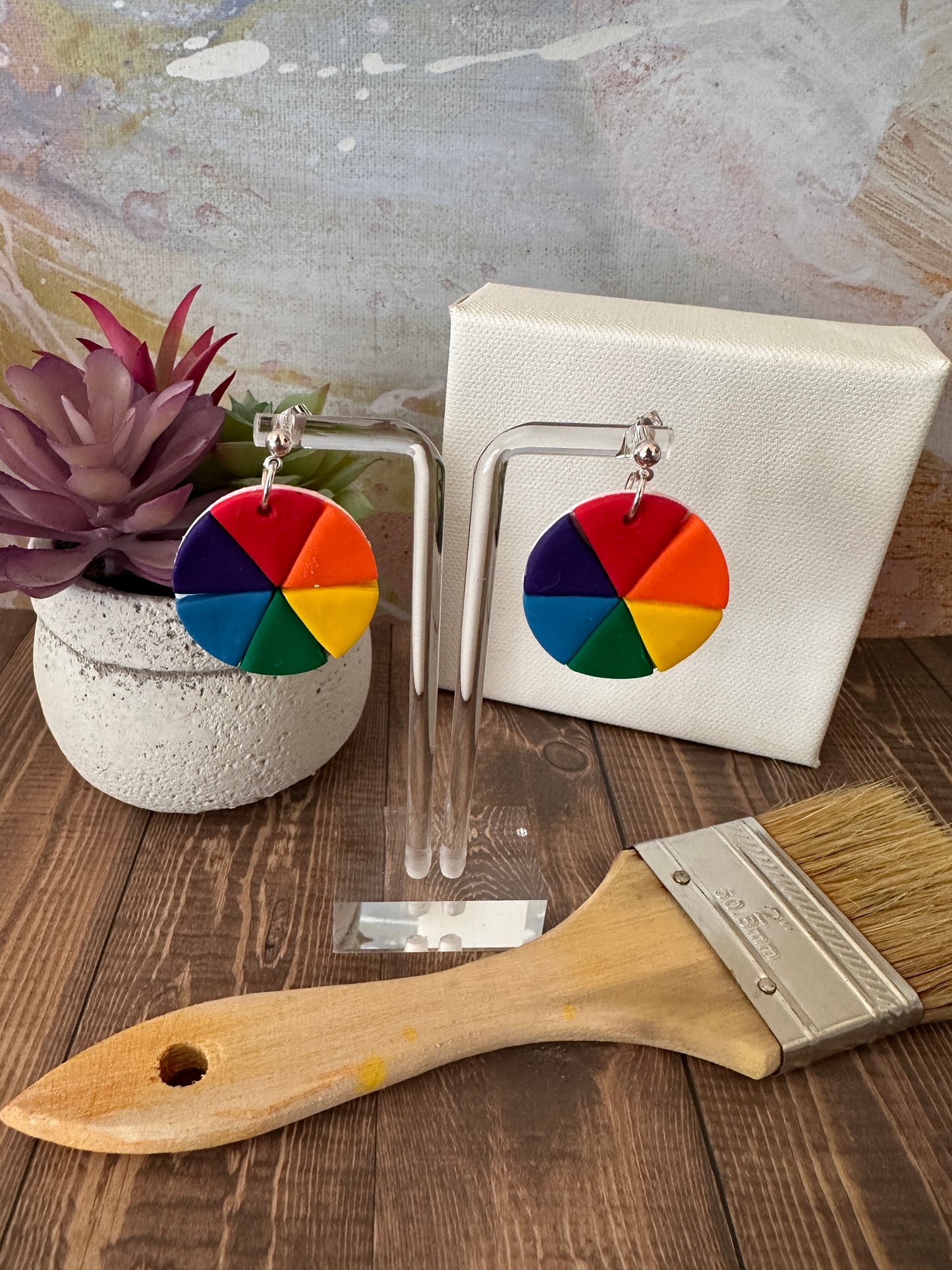 Handmade color wheel polymer clay earrings hanging on an earring stand with a mini canvas, paintbrush, and potted plant in the background.