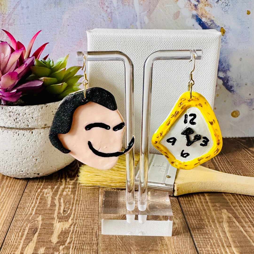 Detailed close up of large Dali earrings showing black hair and handpainted long mustache along with a melted clock with handpainted numbers hanging on an acrylic earring stand with a canvas background.  