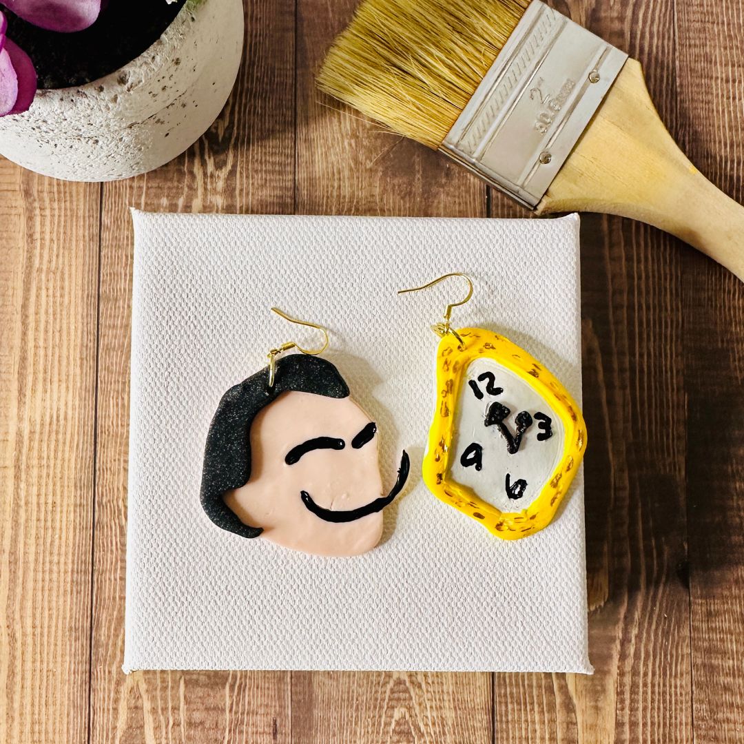 Surreal Dali earrings on a small white canvas with a paintbrush and plant in the background on a wooden surface.