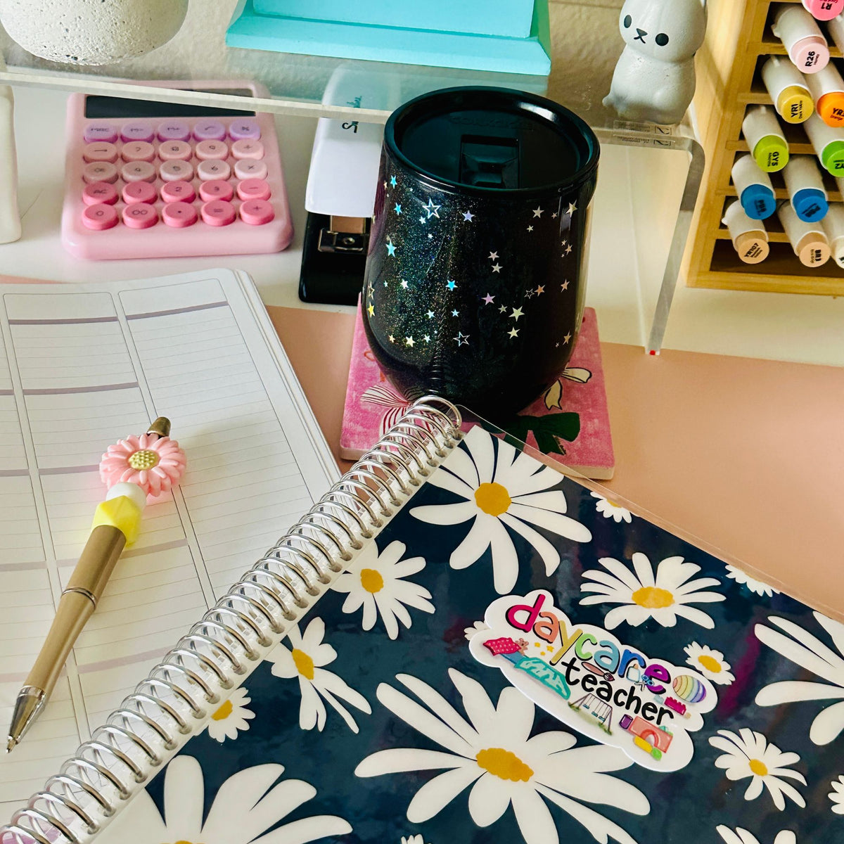 A styled desk scene featuring a navy blue spiral notebook with a white daisy floral pattern and a colorful 'daycare teacher' sticker, alongside a black glittery cup, a pink patterned pen, and other stationery items.