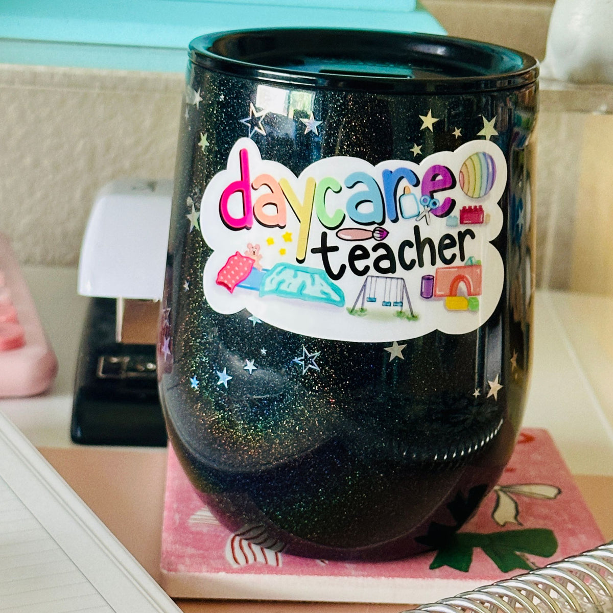 A black glitter stemless wine tumbler with a colorful 'daycare teacher' sticker on the front, resting on a pink patterned coaster on a desk.