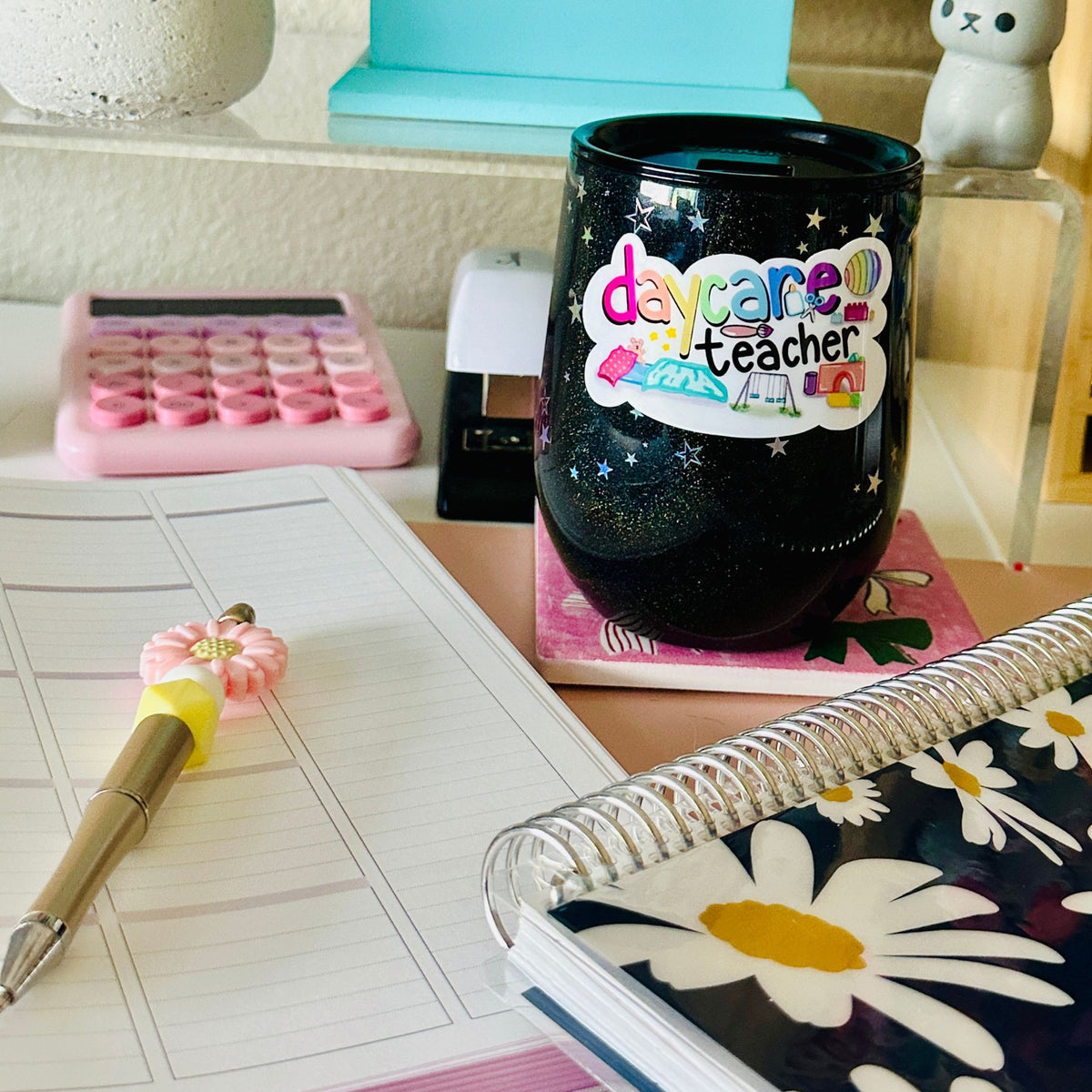A black glitter tumbler cup with a colorful 'daycare teacher' sticker sits on a desk surrounded by school supplies, including a pink calculator, a floral pen, a notebook, and a spiral planner.