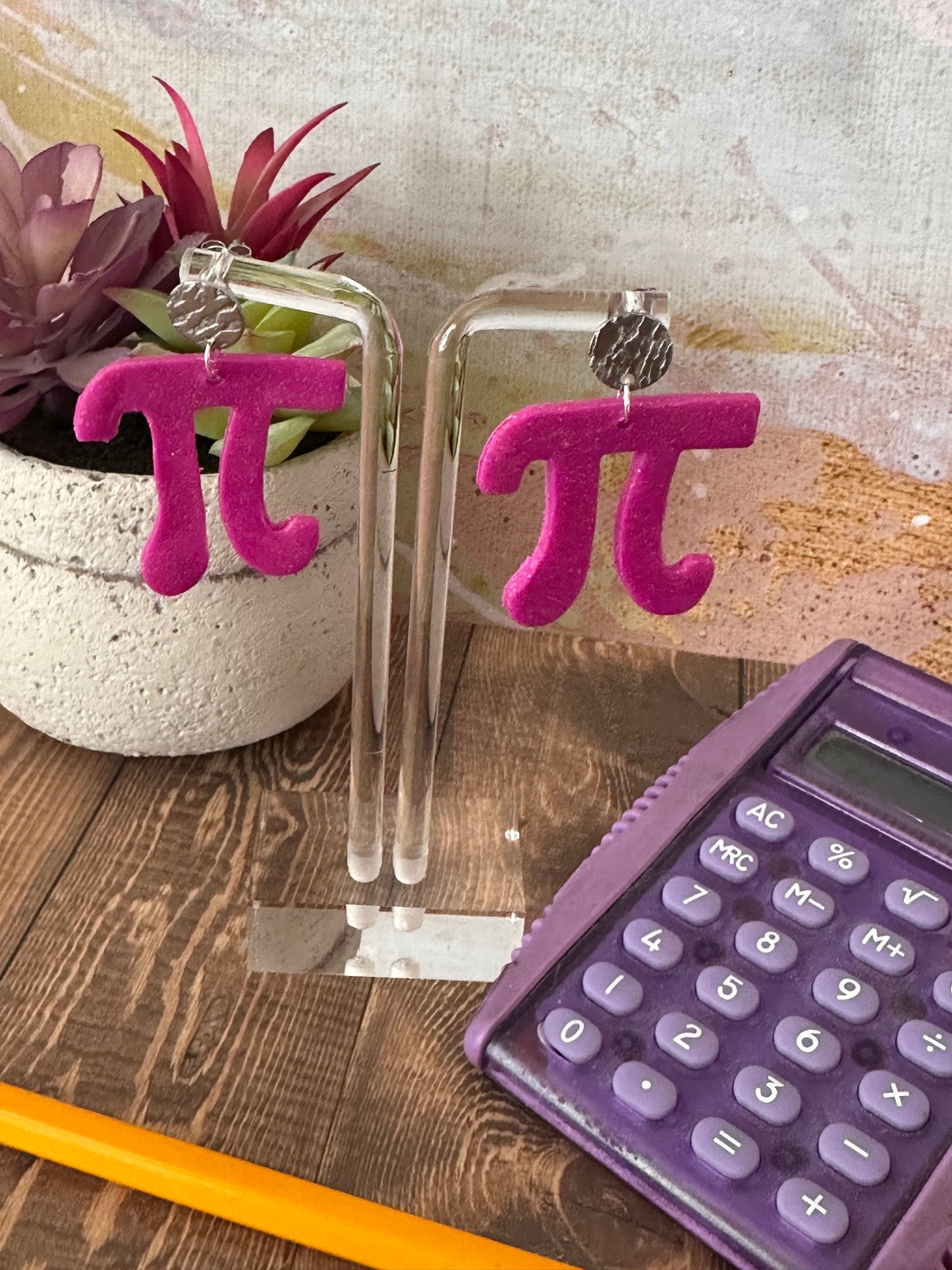 Display of a pair of pink 'π' symbol earrings, purple calculator, and yellow pencil on a wooden surface.