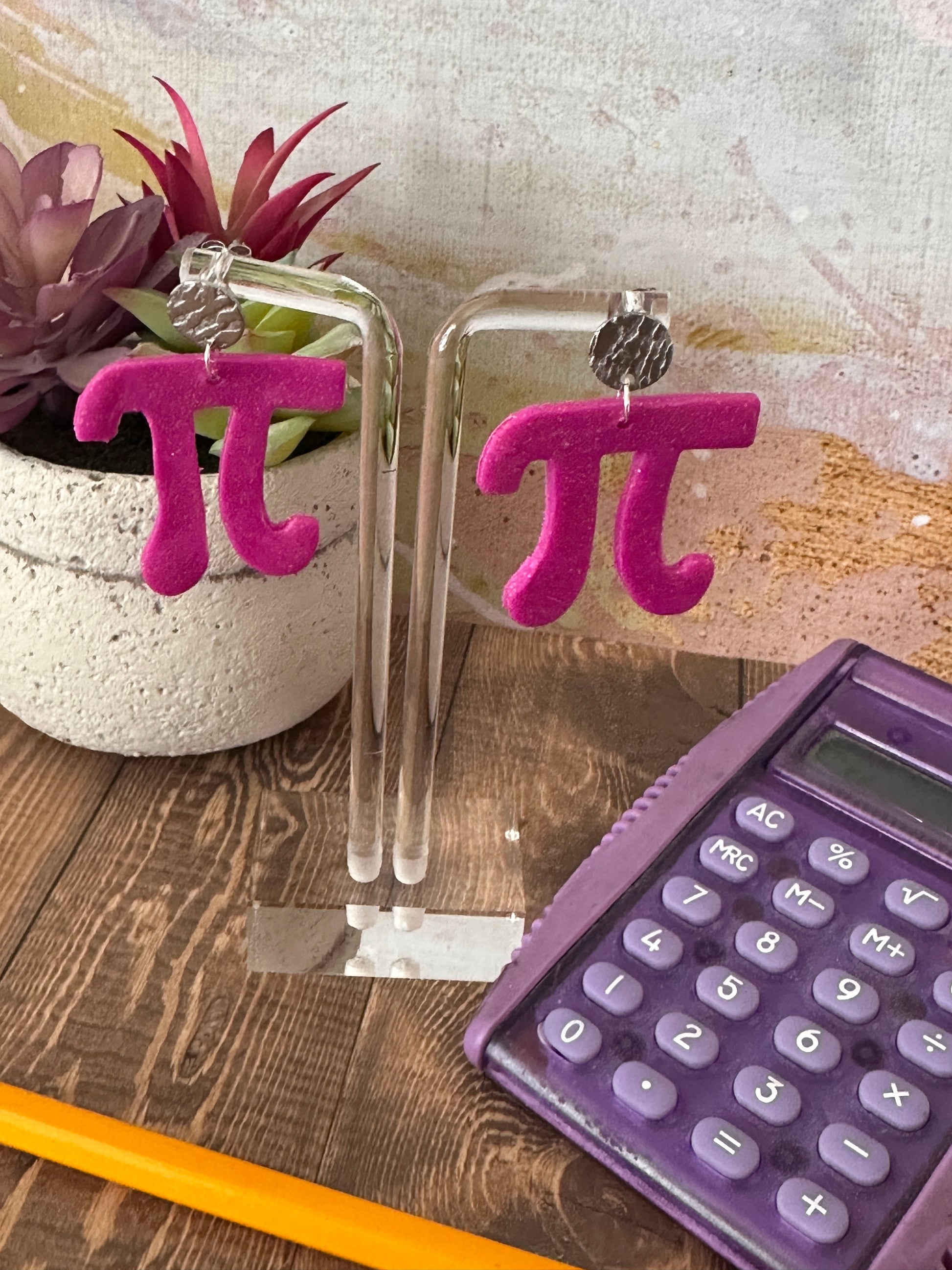 Display of a pair of pink 'π' symbol earrings, purple calculator, and yellow pencil on a wooden surface.