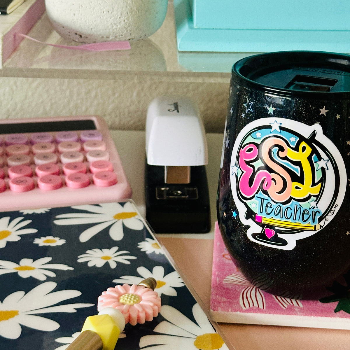 A black insulated tumbler with a colorful 'ESL Teacher' sticker sits on a desk next to a pink floral notebook, a pink calculator, and a white stapler.