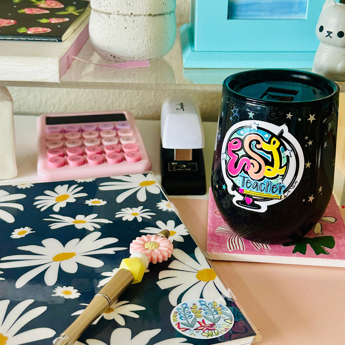 A black glittery insulated tumbler cup with a colorful 'ESL Teacher' sticker, styled on a desk alongside a floral notebook, pink calculator, and other stationery items.