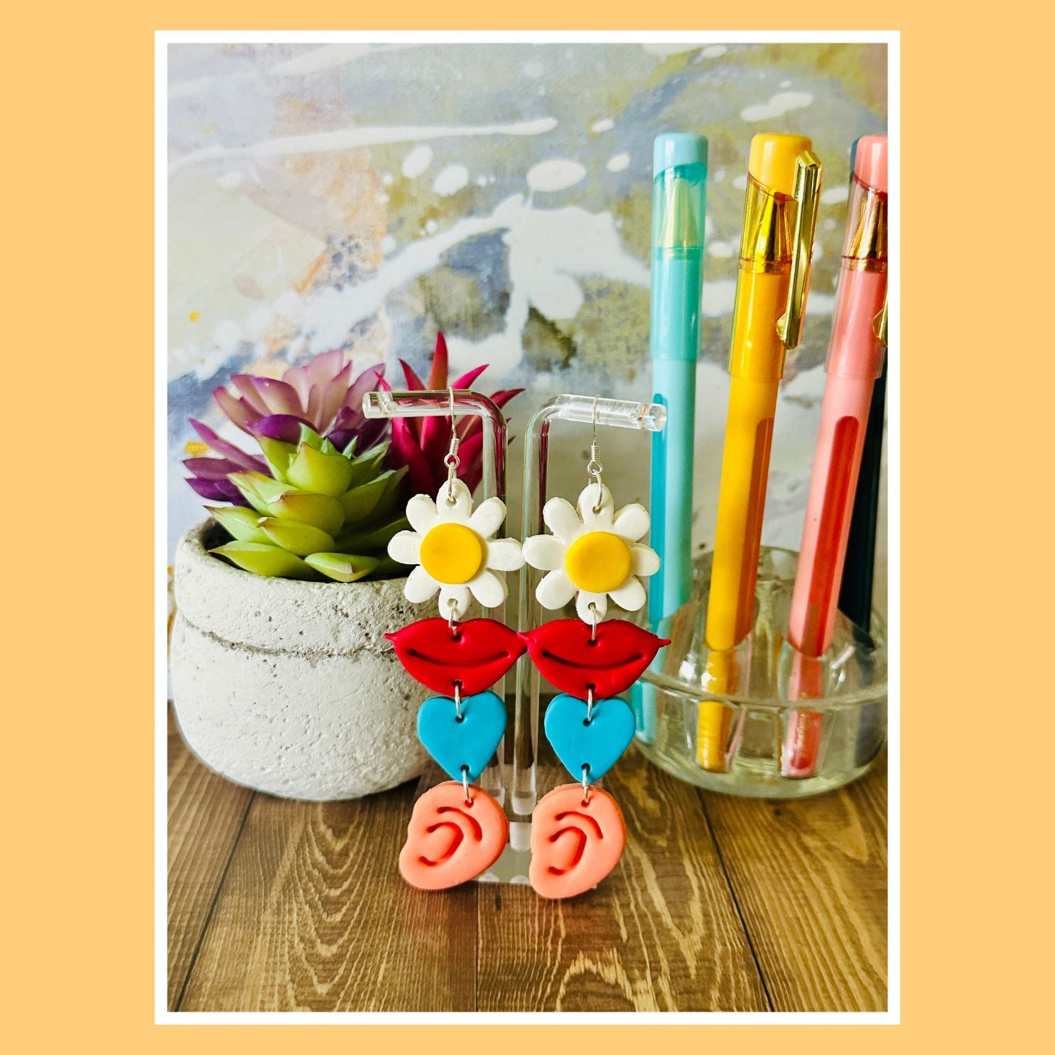 Colorful earrings on a stand with pens and a plant in the background