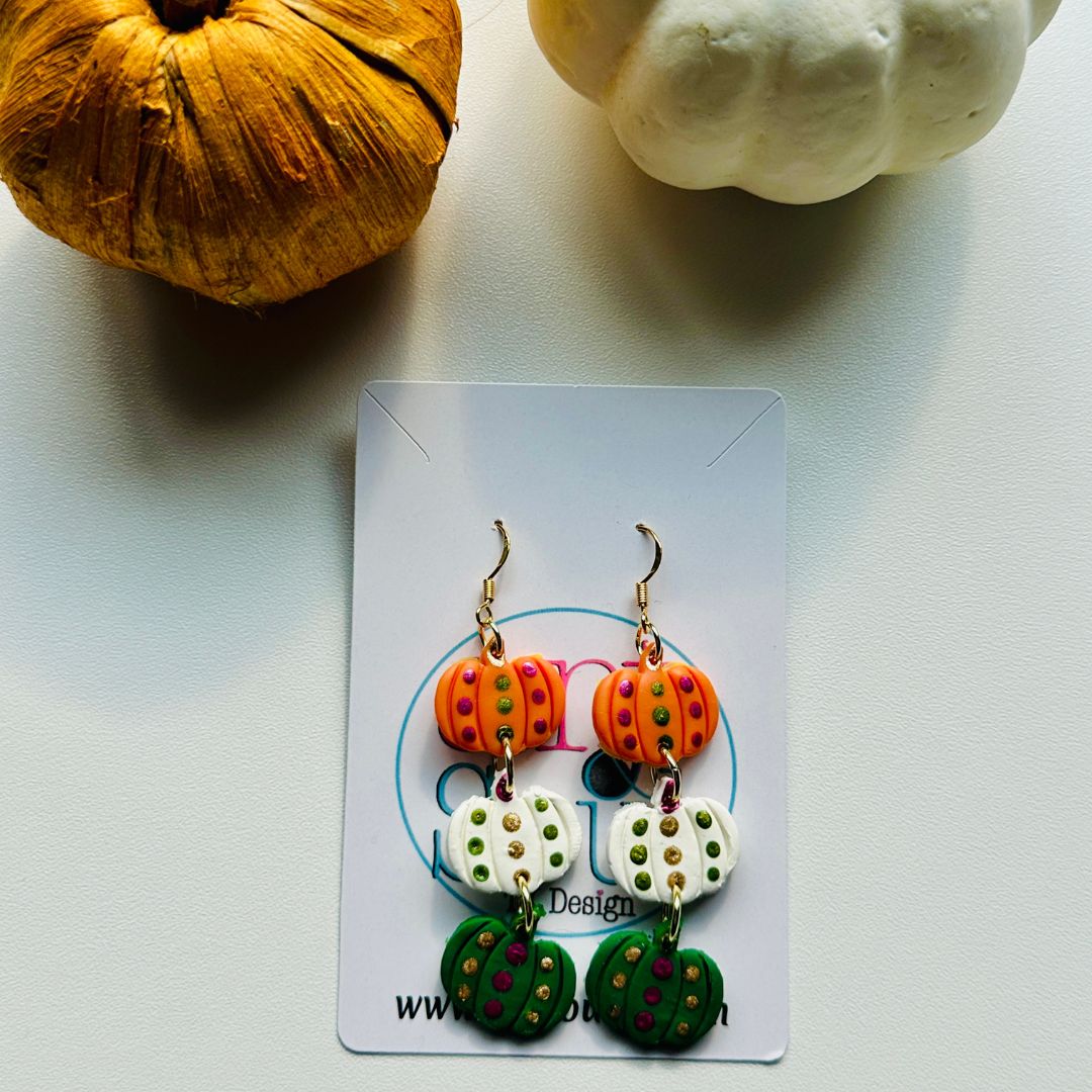 Handmade polka dot pumpkin polymer clay earrings laying flat on a white surface with decorative pumpkins, showing orange, white, and green pumpkin charms with colorful dot details