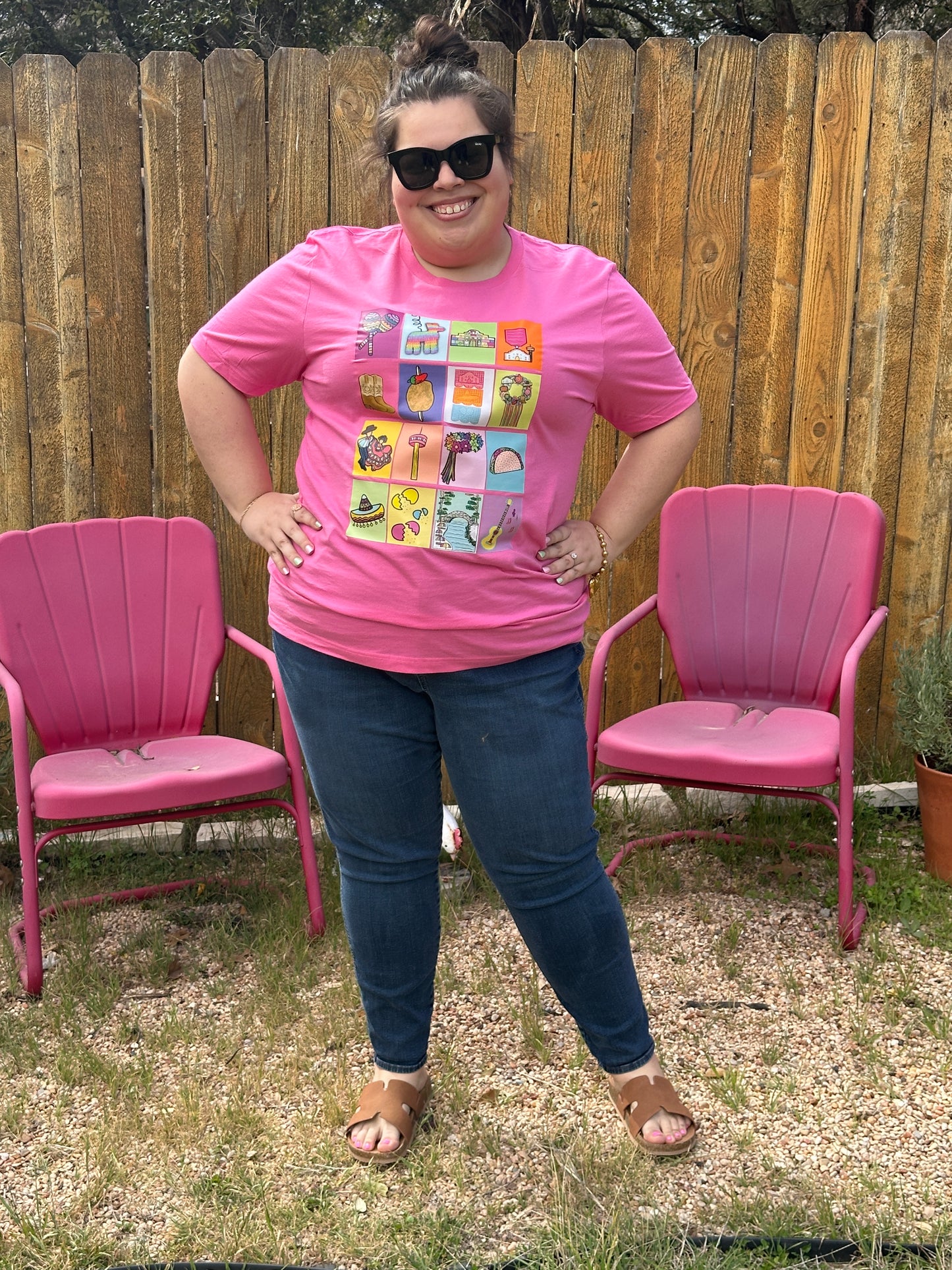 Full body shot of woman in sunglasses and blue jeans wearing a pink Fiesta San Antonio Lotería shirt. 