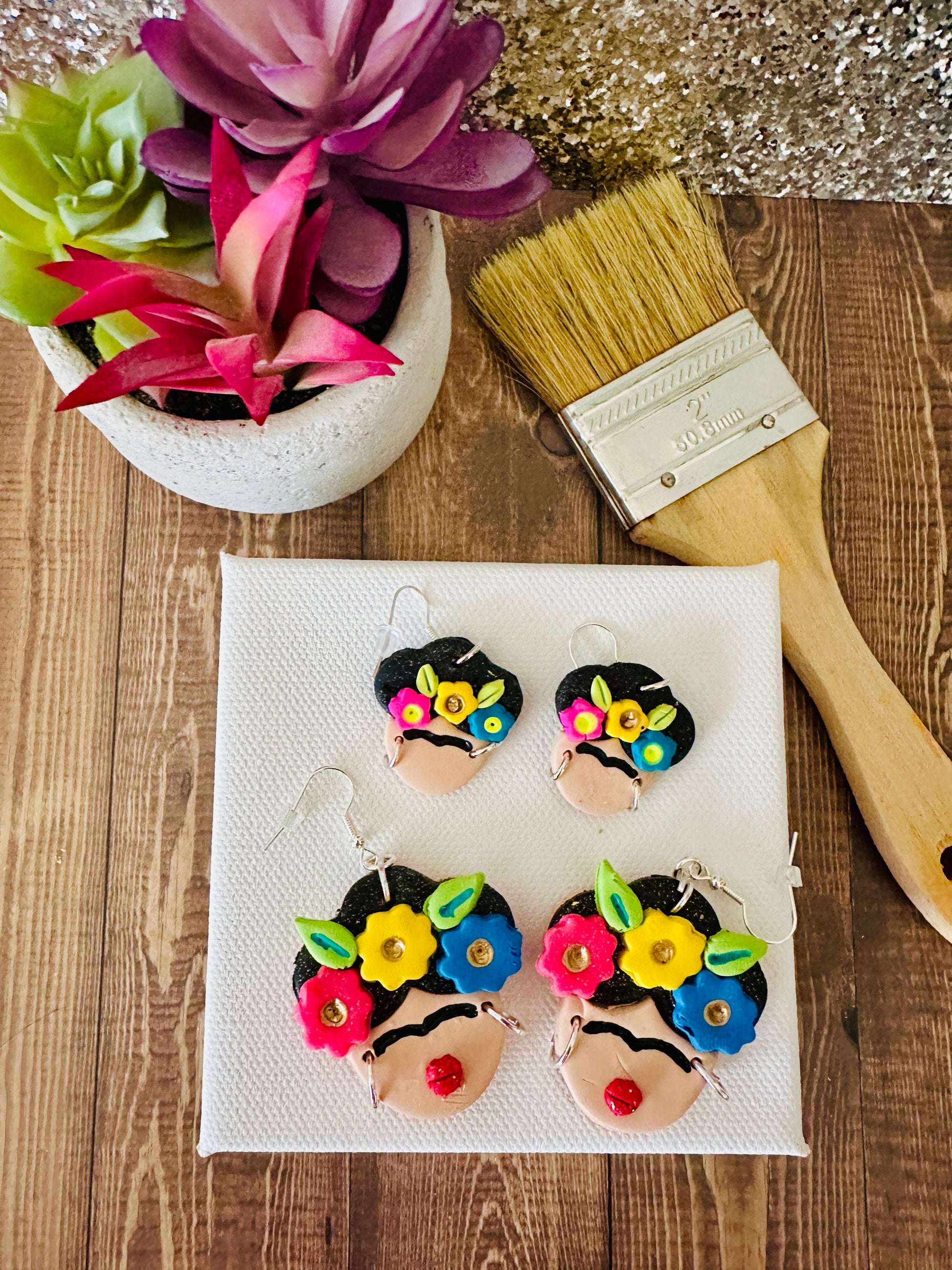 Large and small variants of Frida Kahlo-themed earrings on a white canvas with a brush and plant in the background.