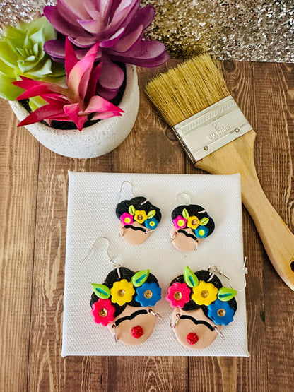 Large and small variants of Frida Kahlo-themed earrings on a white canvas with a brush and plant in the background.