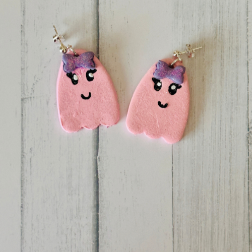 Handmade friendly pink ghost polymer clay earrings on white wooden table with bowties, hand-drawn details, Halloween teacher.