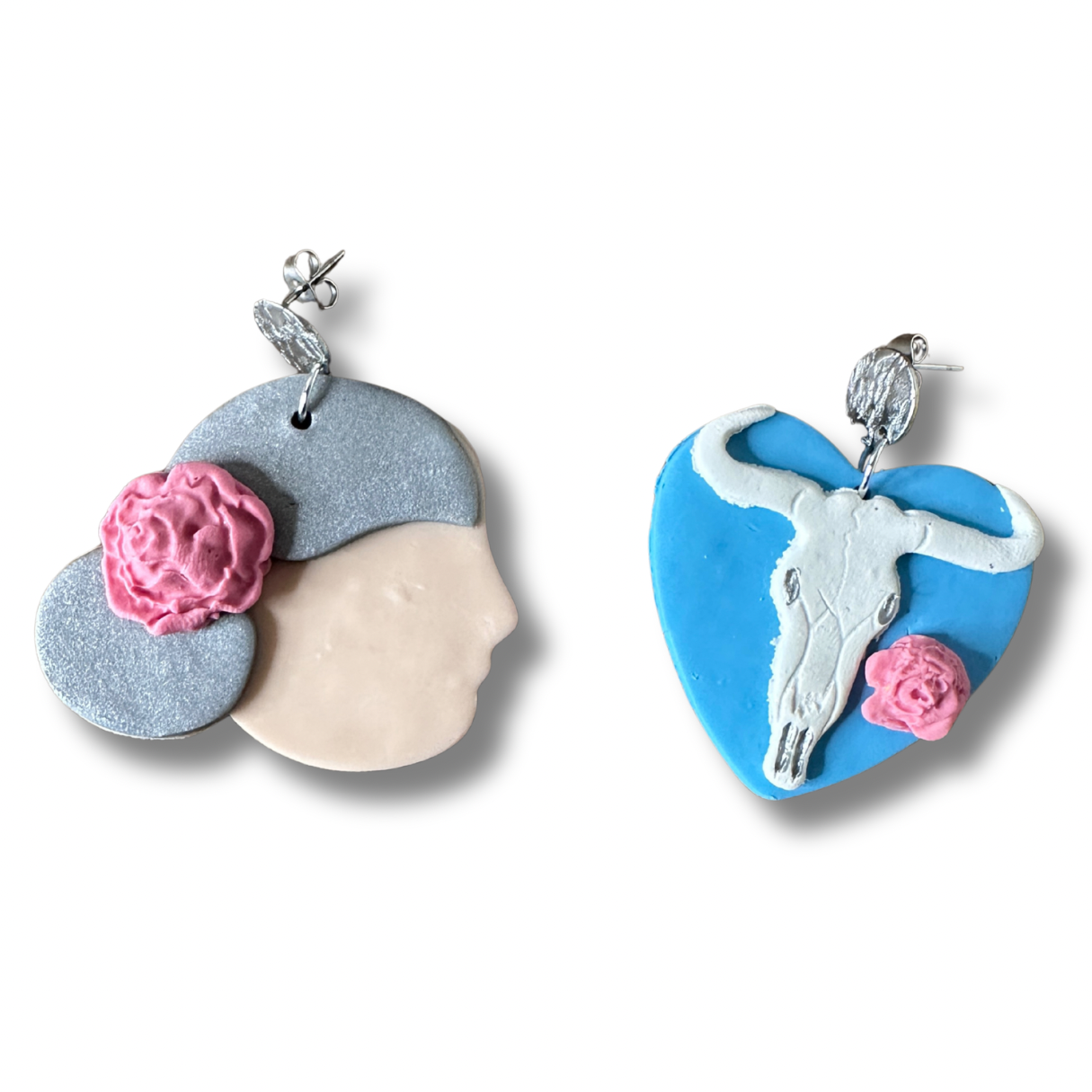 andmade Georgia O'Keeffe polymer clay earrings — left earring is a side profile portrait, right earring is a cow skull and flower on a blue heart, on a white background.