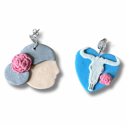 andmade Georgia O'Keeffe polymer clay earrings — left earring is a side profile portrait, right earring is a cow skull and flower on a blue heart, on a white background.