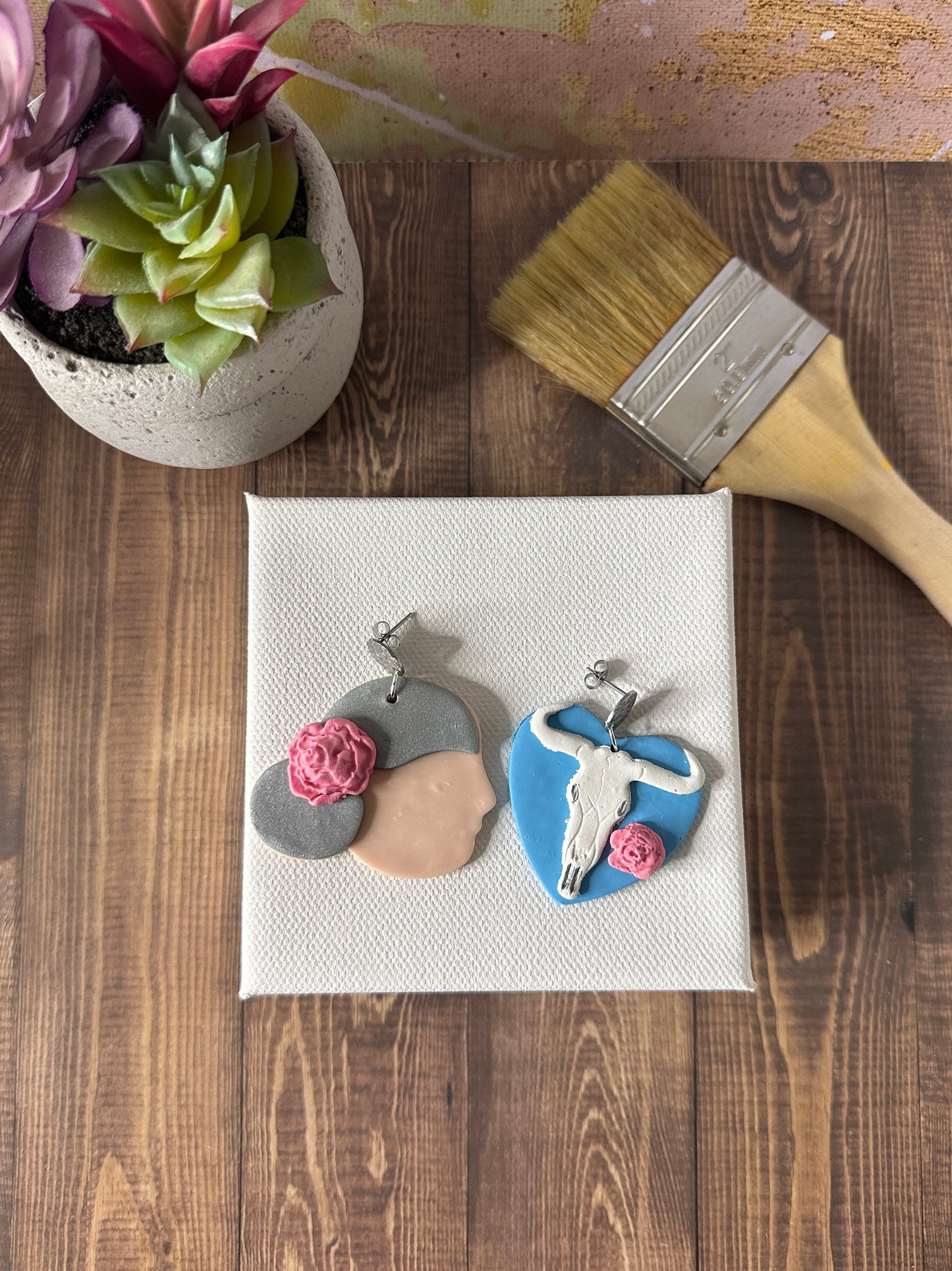 Georgia O'Keeffe handmade earrings resting on a small white painter's canvas with paintbrush and flower pot nearby.