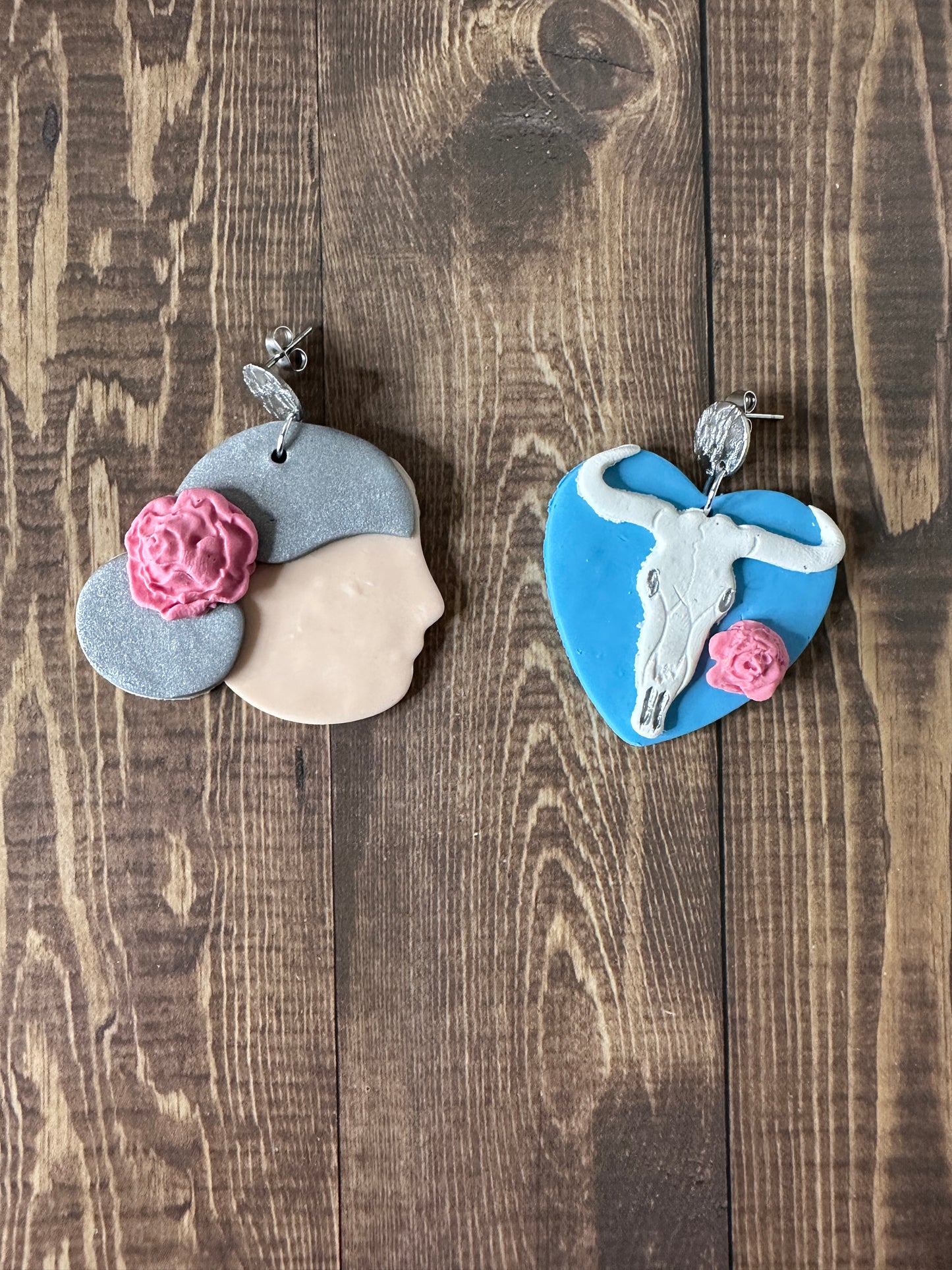 Handmade Georgia O'Keeffe polymer clay earrings displayed on a wooden table, showing portrait and cow skull flower design.