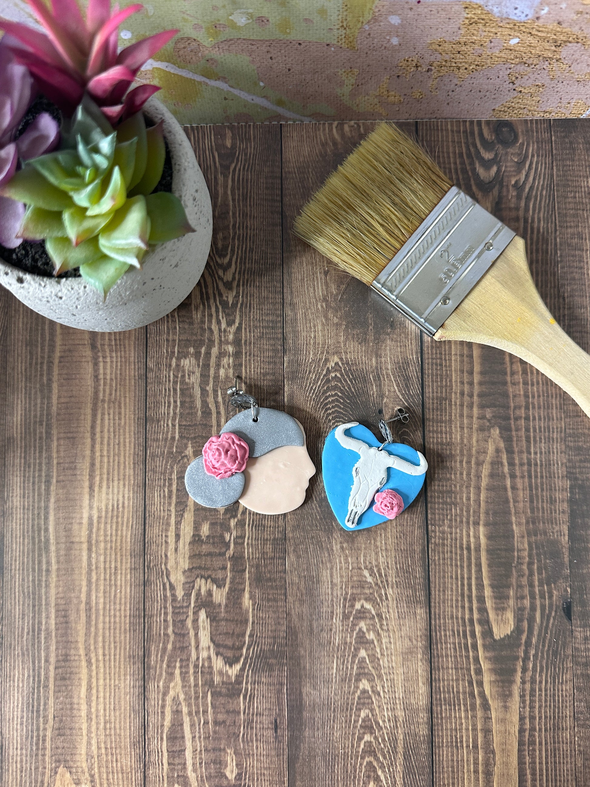 Georgia O'Keeffe polymer clay earrings styled on a wooden table next to a paintbrush and flower pot.