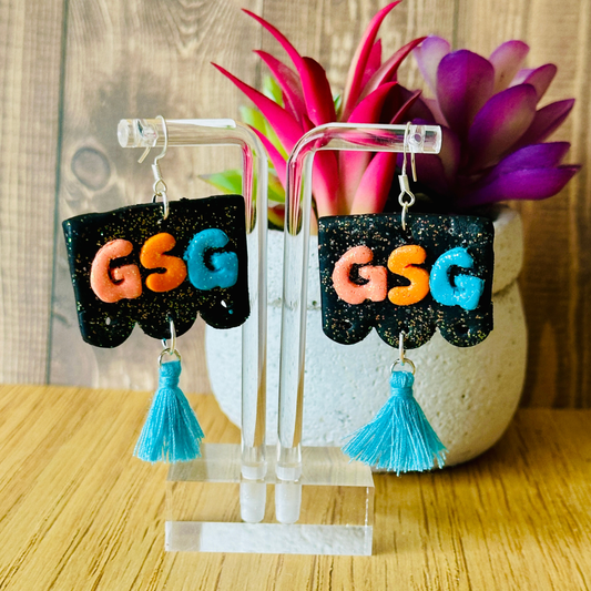 Handmade black GSG papel picado polymer clay earrings with blue tassel on clear earring stand, sterling silver hooks.