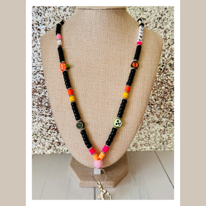 Detailed view of Halloween lanyard with vibrant multicolored beads in orange, purple, black, and green arranged in decorative pattern, displayed on tan mannequin bust against textured gray wall, highlighting the individual bead details, Halloween charms, and sturdy silver metal clasp attachment.