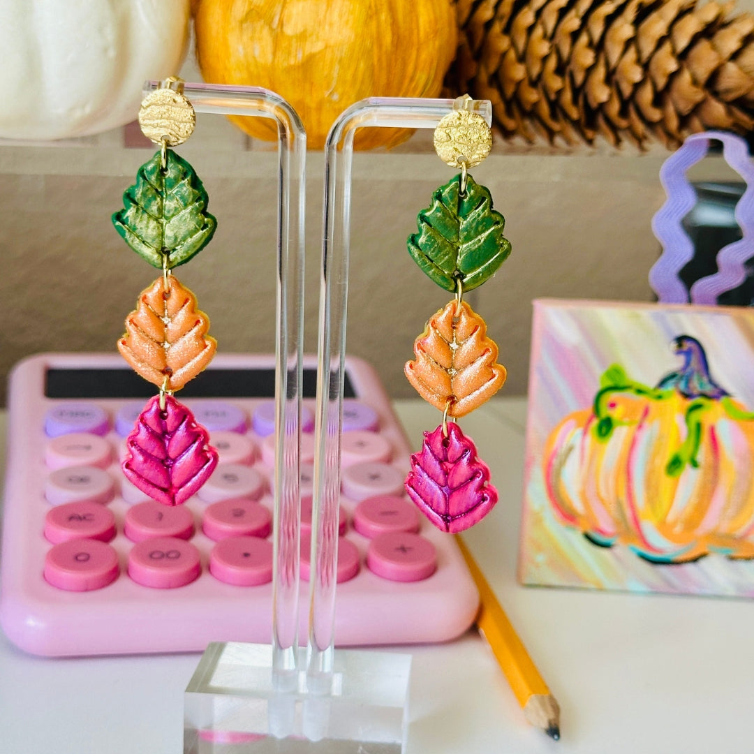 A pair of dangling earrings displayed on a clear acrylic stand on a tabletop. Each earring has three stacked leaf-shaped charms in forest green, pumpkin orange, and cranberry, with a small gold bronze earring post at the top.