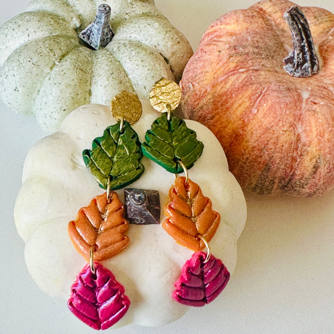 A pair of handmade autumn-themed drop earrings displayed on a white decorative pumpkin and two pumpkins, orange and light green in the background.