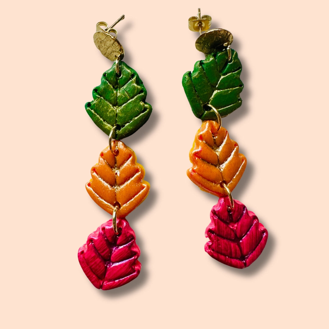 A pair of dangling earrings with three stacked leaf-shaped charms in forest green, pumpkin orange, and cranberry, attached to gold-tone posts, shown against a solid pale peach background.