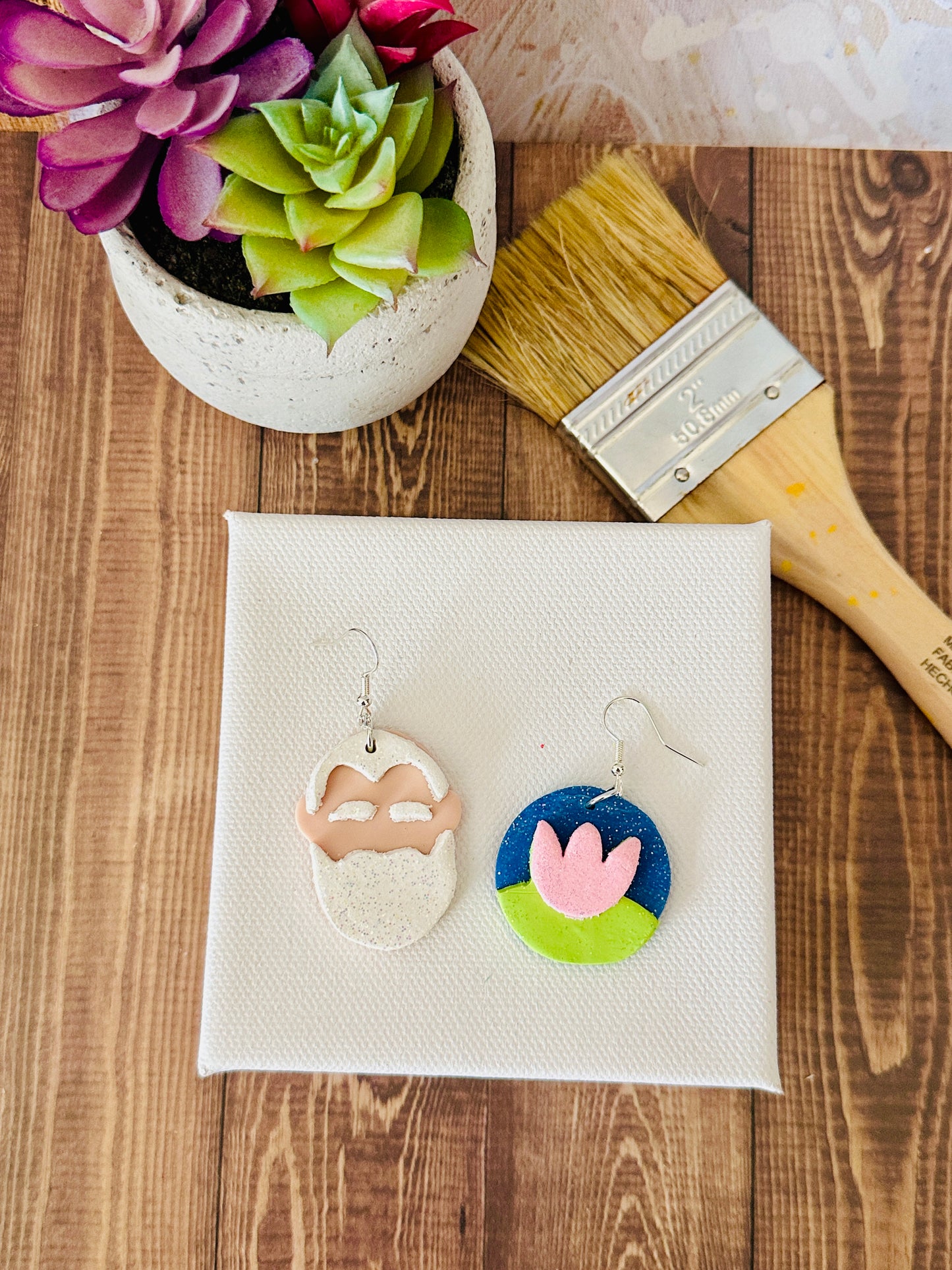 Polymer clay Claude Monet Impressionist earrings on white canvas with paintbrush and plants, different angle view.