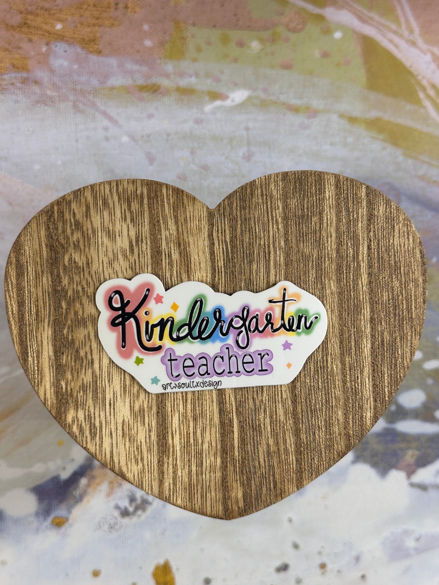 Kindergarten teacher vinyl sticker on a wooden heart-shaped table with colorful background, showing bold cute font with colorful letter highlights.