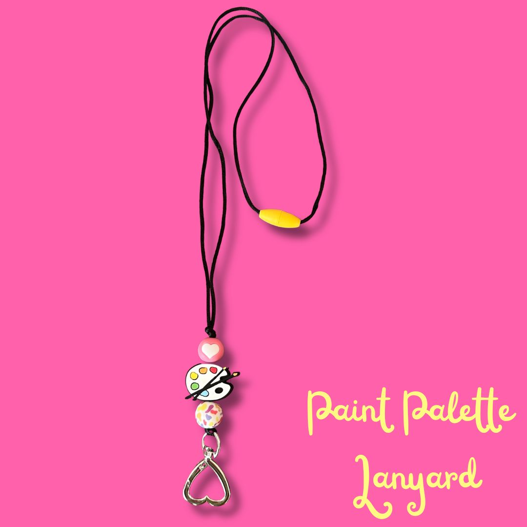 A paint palette themed cord lanyard necklace featuring a black cord, a yellow breakaway clasp, silicone beads, and a silver heart-shaped ring, set against a solid pink background with yellow text reading 'Paint Palette Lanyard'.