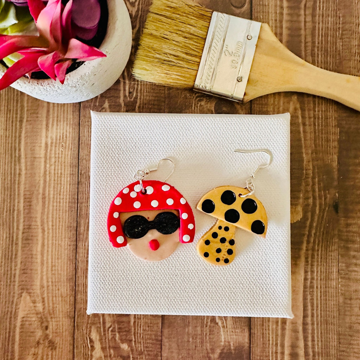 Large Yayoi Kusama polymer clay earrings with detailed portrait, red polka dot hair, black sunglasses and yellow mushroom on a blank canvas and wooden table