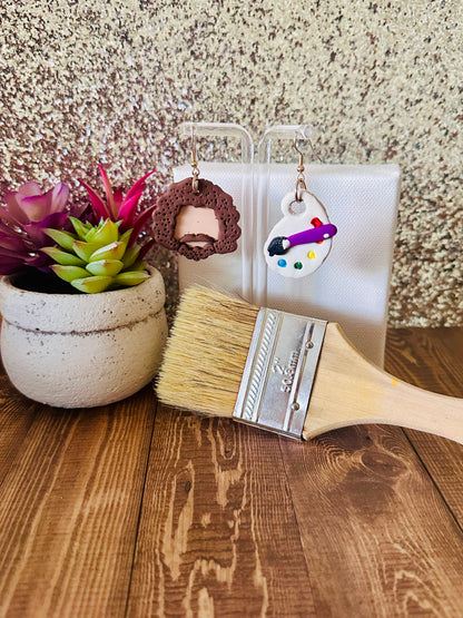 Large Bob Ross earring set displayed by a paint canvas with a paintbrush and a plant on a wooden surface.