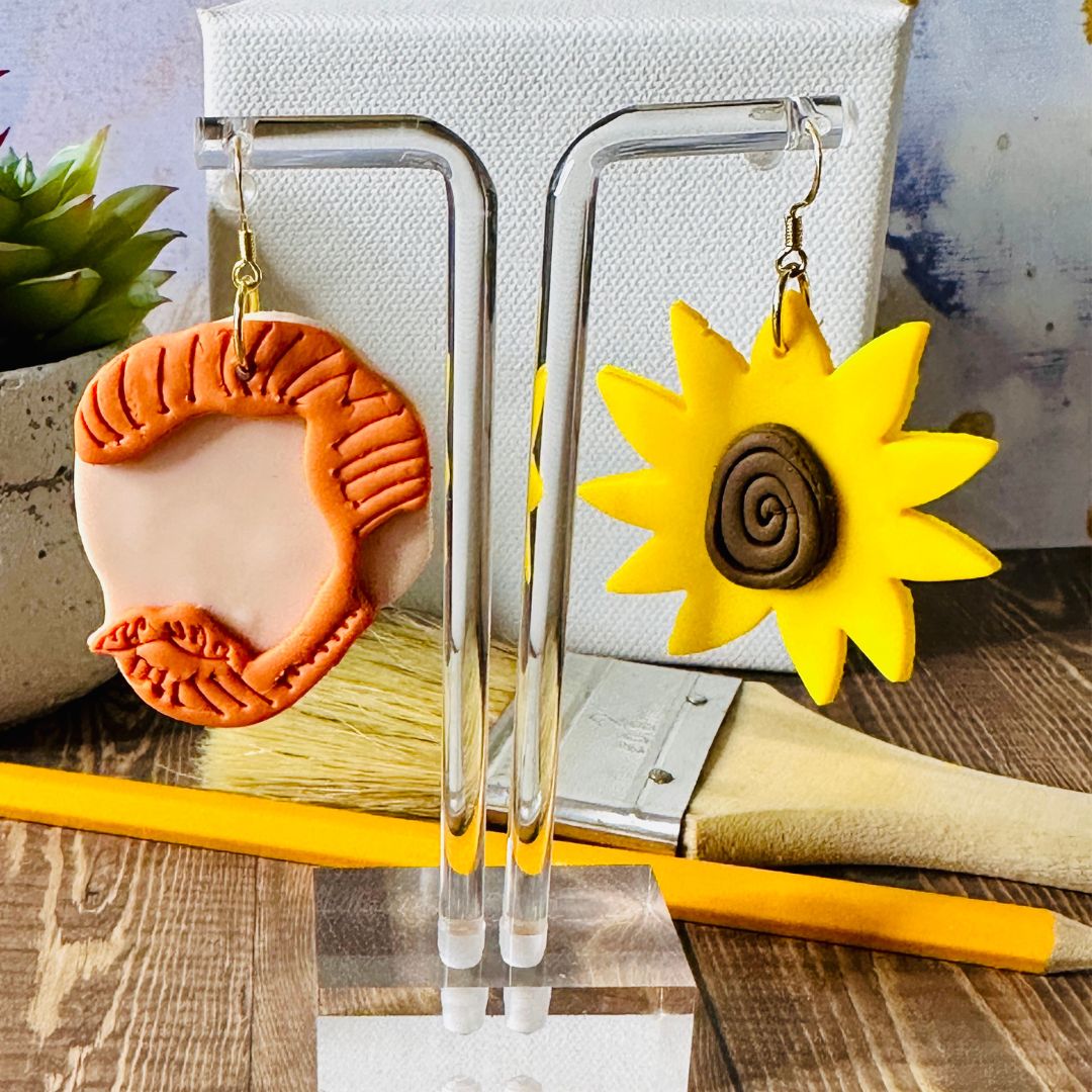 Detailed close up of large Van Gogh earrings showing red har texture and sunflower swirls on a clear stand with a white canvas background. It's resting on a wooden base with a paintbrush and pencil on top. 