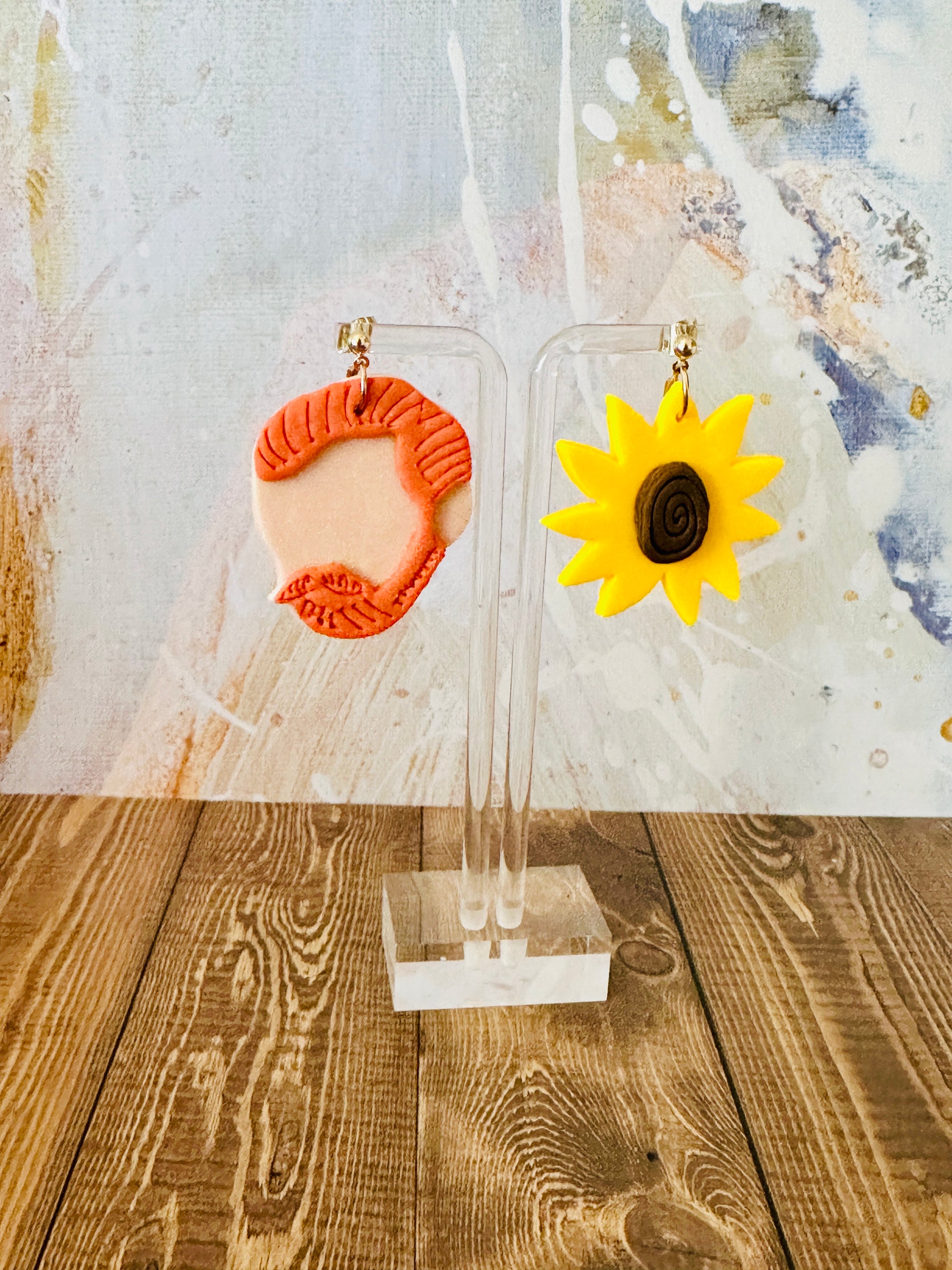 Close up of large handmade Van Gogh sunflower earrings on display stand with wooden table and colorful abstract backdrop.