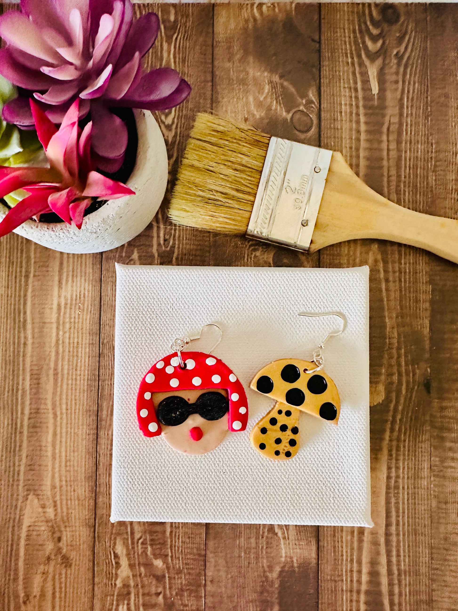 Large Yayoi Kusama polymer clay earrings with detailed portrait, red polka dot hair, black sunglasses and yellow mushroom on a blank canvas and wooden table