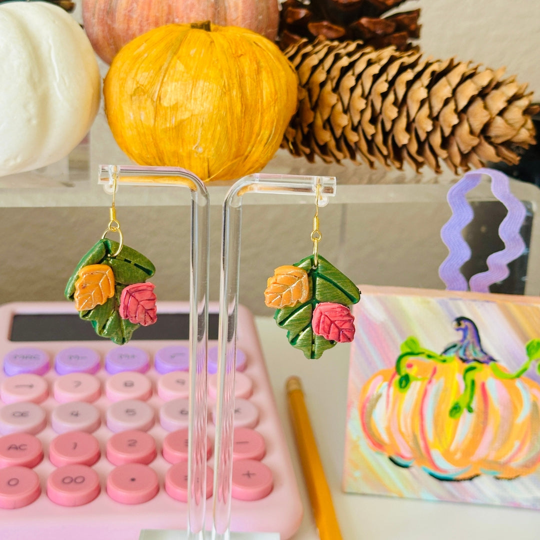A pair of colorful autumn-themed dangle earrings displayed on a clear acrylic stand. The earrings feature forest green leaf shapes with small pumpkin orange and cranberry leaf details, hanging from gold-toned hooks.