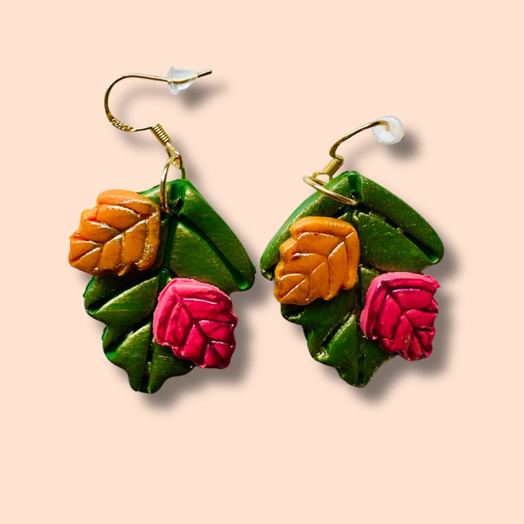 A pair of handmade polymer clay earrings shaped like autumn leaves, featuring textured forest green leaves with pumpkin orange and cranberry leaf accents, suspended from gold-tone fishhook wires on a solid pale peach background.