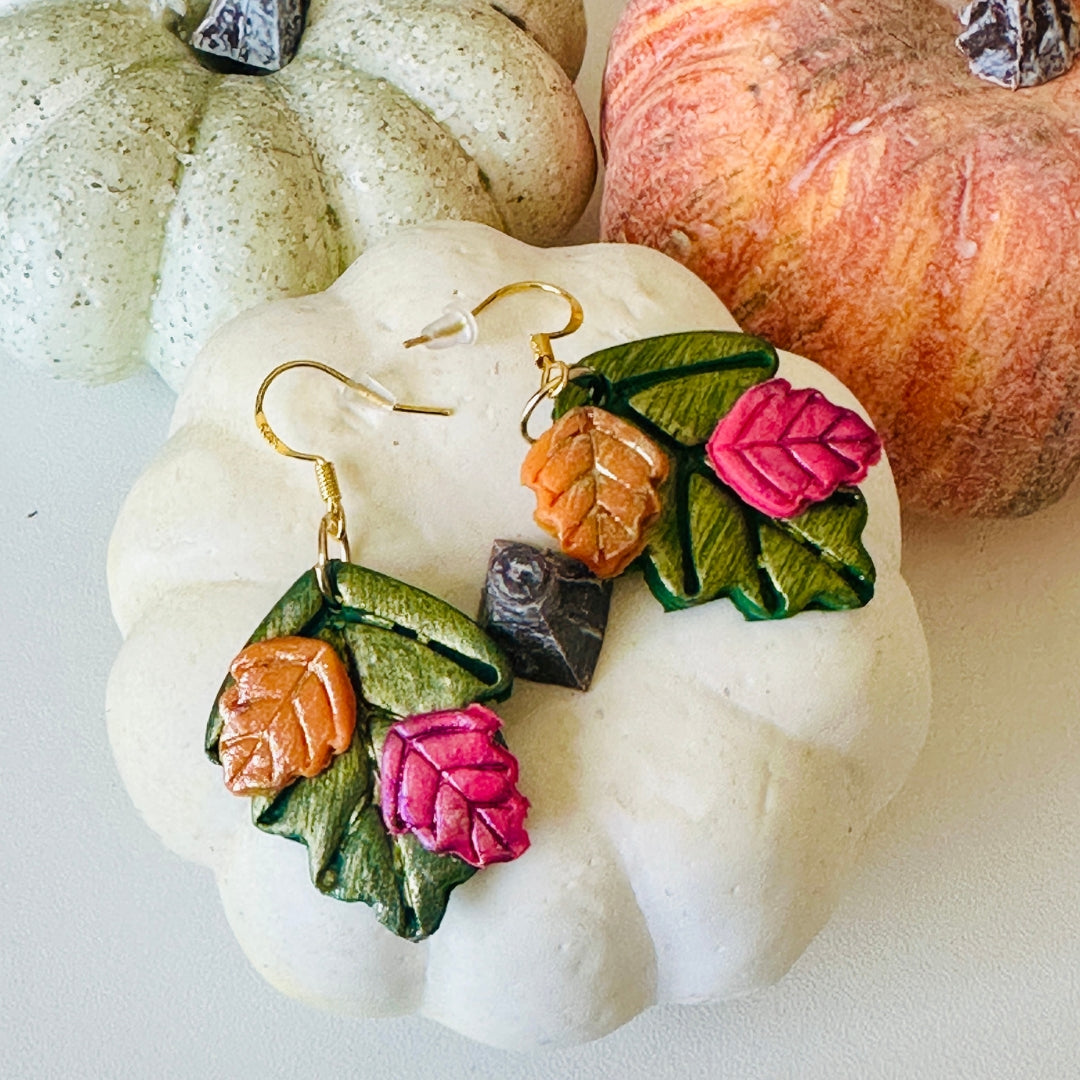 Autumn-themed drop earrings with forest green leaves, small cranberry and pumpkin orange leaf accents, and gold fishhook wires, displayed on a white decorative pumpkin with other pumpkins in the background.