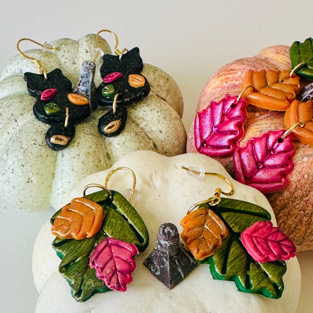 Three pairs of handmade polymer clay earrings displayed on decorative fall pumpkins. The pair on the left features a black cat silhouette with colorful fall leaves and a gold wire earring hooks, on the right features dangled Harvest Drop earrings and a gold wire earring hooks, and at the bottom center the featured product, Leaf Bouquet earrings on a white pumpkin.