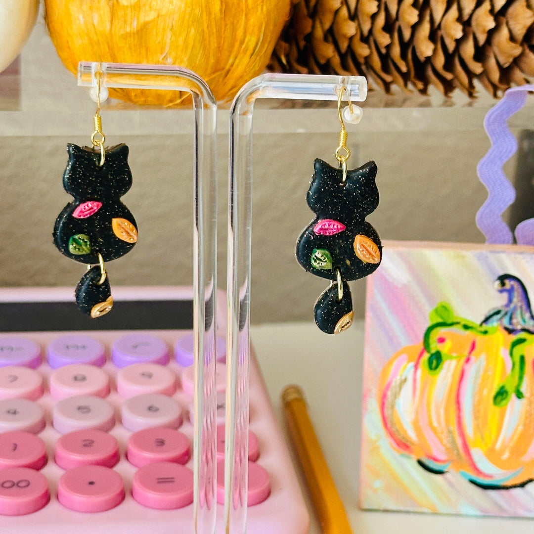 A pair of black glitter cat-shaped dangle earrings with colorful painted leaf accents, displayed on a clear acrylic stand in a styled tabletop setting with pumpkins, a pink calculator, and a small fall pumpkin painting.