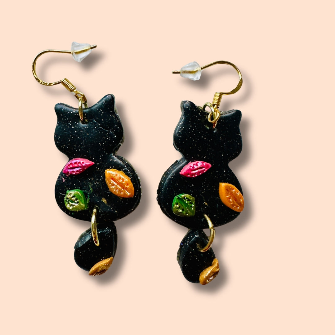 A pair of black cat-shaped drop earrings with gold fishhook wires and colorful raised details, shown on a solid peach background.