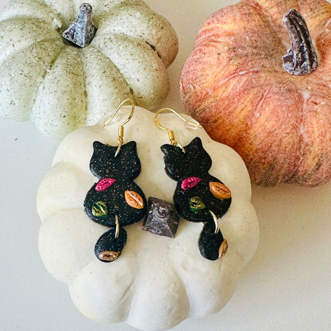 Black cat-shaped drop earrings with colorful leaf details and gold fishhook wires, displayed on a white decorative pumpkin with two small pumpkins in the background.