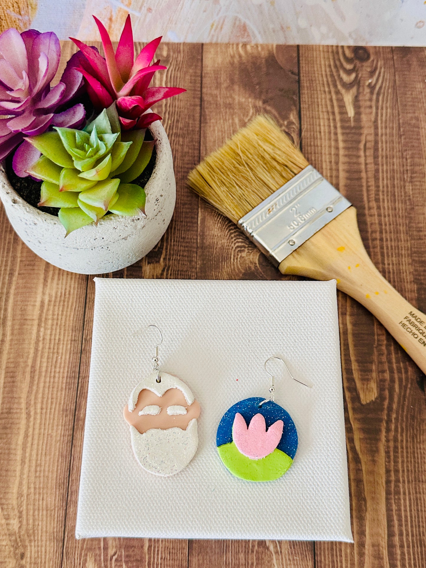 Claude Monet artist earrings laying flat on white canvas surrounded by paintbrush and potted plants on wooden table.