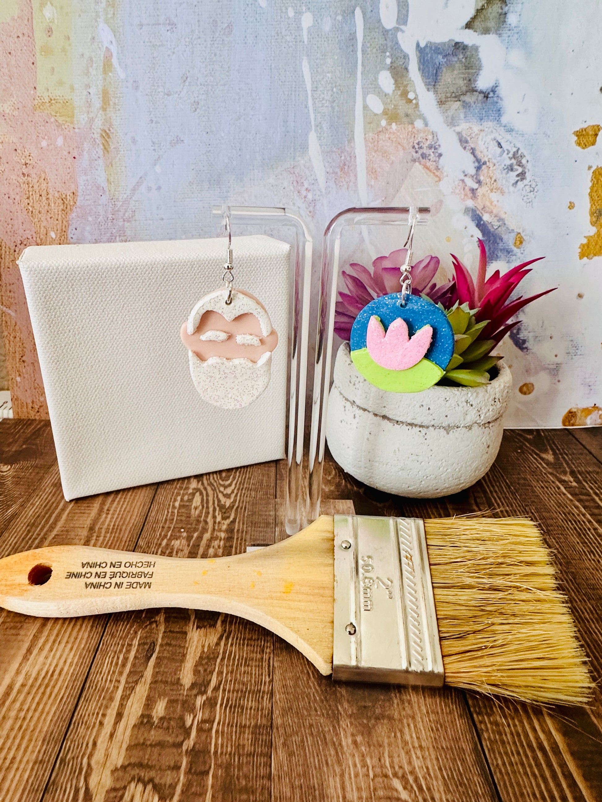 Art teacher gift Monet earrings on stand with canvas, angled paintbrush, potted plants on wood table, abstract wall.