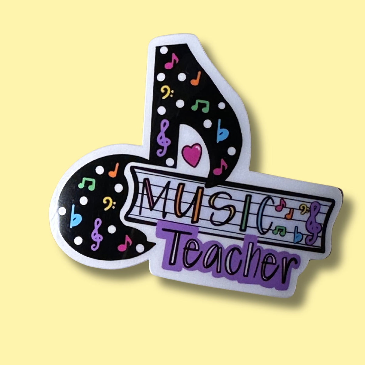 A die-cut sticker shaped like a musical note with the words 'MUSIC Teacher' on a light yellow background. The sticker has a black base decorated with colorful musical notes, treble clefs, dots, and a pink heart, with 'MUSIC Teacher' written across it in stylized multicolored lettering.