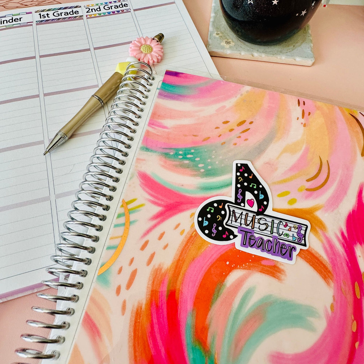 A colorful spiral-bound planner or notebook cover with a pink, orange, and teal abstract swirl pattern, featuring a sticker that reads 'Music Teacher.' A gold pen with a pink flower charm rests on an open lined weekly planner page, with a dark starry mug in the background on a peach tabletop.