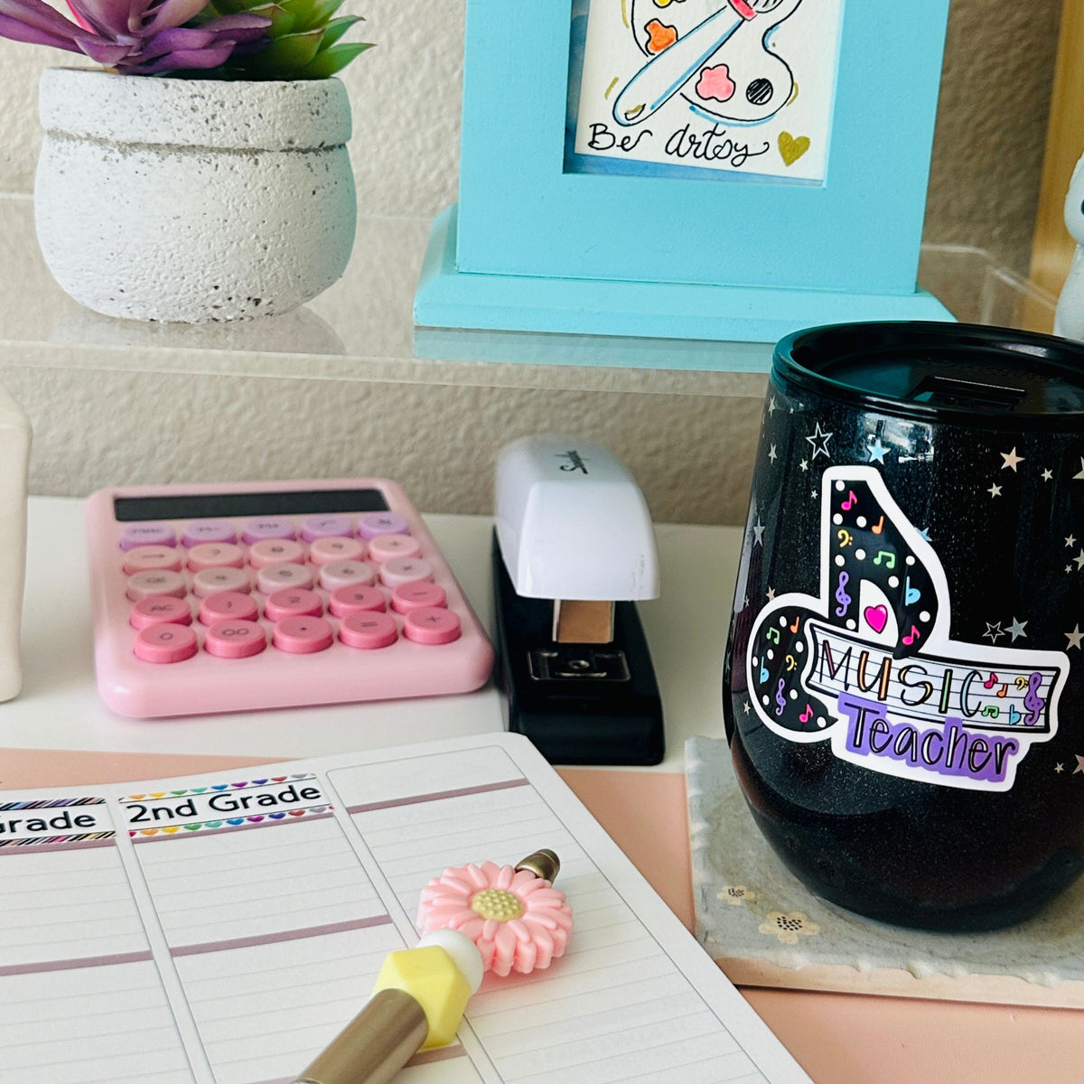 A black glitter tumbler cup with a 'Music Teacher' sticker sits on a desk next to a pink calculator, white stapler, planner, and small potted succulent.